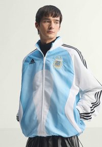 Νεαρός άνδρας φορώντας ανοιχτό γαλάζιο και λευκό μπουφάν ποδοσφαίρου Αργεντινής με το λογότυπο της Adidas, στέκεται με τα χέρια στις τσέπες, κοιτάζοντας πλάγια.