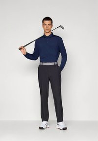 Golfantrekk med en marineblå langermet polo, grått belte, mørke bukser og hvite golfsko, med en golfklubbe i hånden.