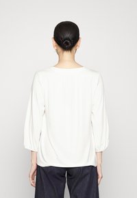 Camicia bianca leggera con vestibilità rilassata, caratterizzata da maniche a tre quarti e dettaglio posteriore plissettato. Abbinata a pantaloni in denim scuro.