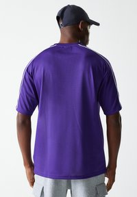Jersey atlético de malla morada con mangas cortas, que presenta rayas blancas de acento a lo largo de los hombros. Textura suave y transpirable visible.