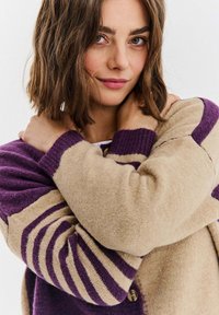 Pletený béžový kardigan s fialovými pruhy, s žebrovanými detaily a knoflíky. Měkká textura a uvolněný střih.