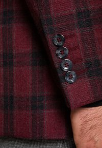 Rękaw bordo marynarki w kratę z czterema ciemnoszarymi guzikami, wykonany z teksturowanej tkaniny z czarnymi akcentami w wzorze.