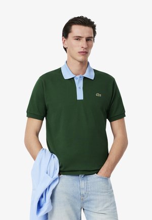 Grön polo skjorta med ljusblå krage, korta ärmar och en liten broderad logotyp på bröstet, kombinerad med ljusblå ytterkläder.