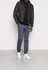 Chaqueta puffer negra con costuras horizontales, superpuesta sobre un hoodie negro. Pantalones de chándal grises y zapatillas blancas completan el atuendo.