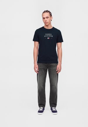 Jeune homme aux cheveux tressés portant un t-shirt noir Tommy Hilfiger, un jean noir délavé et des baskets bleu marine, debout sur un fond uni.
