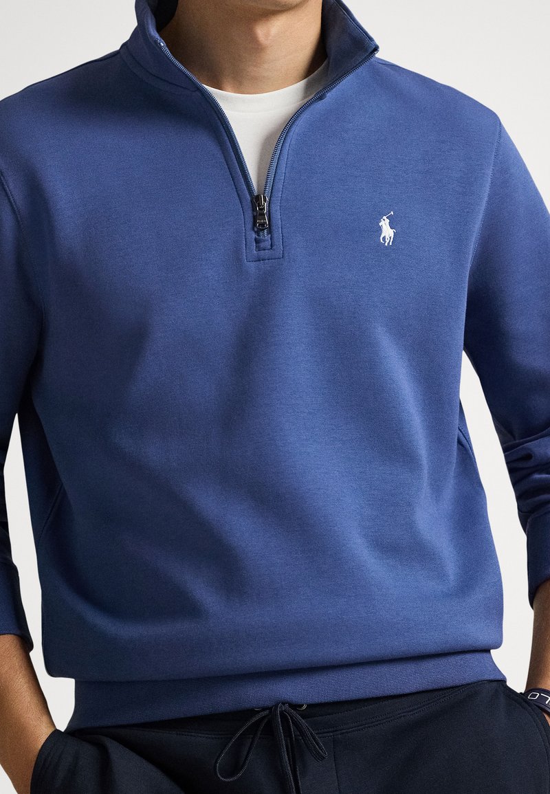 Uomo che indossa una felpa blu con cerniera a quattro quarti e un logo bianco di un giocatore di polo sul petto, abbinata a pantaloni neri con coulisse.