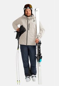 Das Skianzug besteht aus einer hellbeigen Jacke, marineblauen Hosen, schwarzen Handschuhen, weißen Skiern und weißen Skischuhen. Akzente setzen die türkisfarbenen Skibindungen.