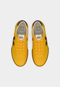 Κίτρινα συνθετικά αθλητικά παπούτσια με μαύρες λεπτομέρειες, με λείο υλικό, στρογγυλεμένη μύτη και επίπεδα κορδόνια. Το λογότυπο «VEJA» είναι εμφανές στη γλώσσα του παπουτσιού.