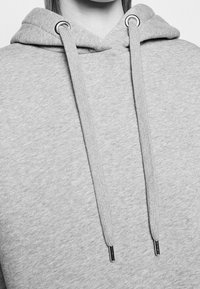 Sudadera gris con cordón, ojales de metal y una tela suave y texturizada. Presenta un bolsillo canguro frontal y un diseño simple.
