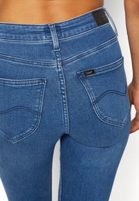 Niebieskie dżinsy z wysokim stanem, dwoma tylnymi kieszeniami i widocznymi szwami. Mała czarna metka z napisem "Lee" na prawej kieszeni.