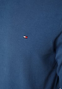 Suéter de malha azul com um pequeno emblema em vermelho, branco e azul na área do peito esquerdo. Textura suave com um design ajustado.