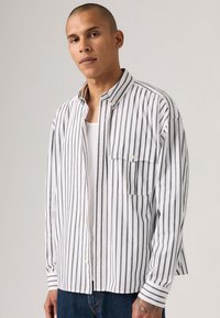 Hvit skjorte med vertikale mørke striper, med en brystlomme og en myk, glatt tekstur. Kombinert med blå jeans.