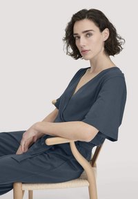 Dunkelblauer Jumpsuit mit tiefem V-Ausschnitt, kurzen Ärmeln und elastischem Bund, kombiniert mit einem Holzstuhl mit geflochtener Sitzfläche.