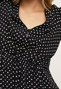 Černé šaty s bílými puntíky. Mají zapínání na knoflíky u výstřihu, nařasený pas a měkkou, strukturovanou tkaninu. Doplňují je detaily v podobě zavinovacího designu.