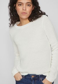 Pulover alb din tricot, cu un model texturat, guler rotund și mansete strâmte, combinat cu blugi blue. Modelul pozează cu mâinile în buzunare.