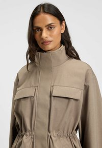 Giacca beige con colletto alto, due tasche sul petto, elastico in vita e texture del tessuto liscia. Dettagli di design minimalisti.