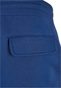 Gros plan sur un pantalon en tissu bleu avec une poche arrière rectangulaire à rabat et des coutures visibles autour de la ceinture.