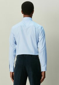 Svetlomodrá kockovaná košeľa s dlhými rukávmi a golierom na gombíky, nosená s tmavými nohavicami, pohľad zozadu.