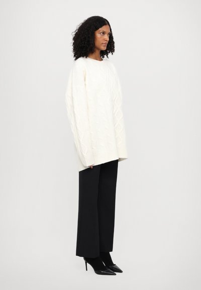 Pull en maille blanche avec des manches surdimensionnées, associé à un pantalon noir large et ajusté et des escarpins noirs pointus. Le modèle pose de profil.