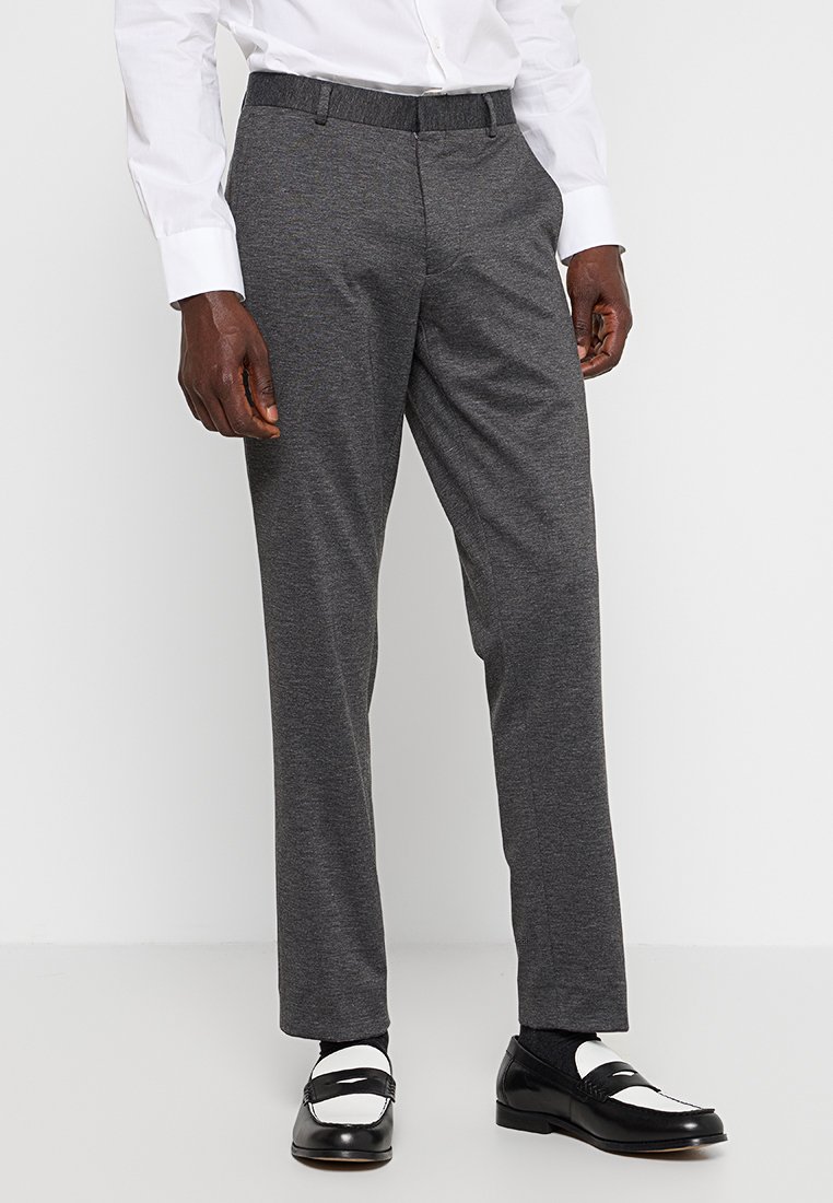 Hombre con pantalones de vestir gris oscuro, mocasines en blanco y negro, calcetines oscuros y una camisa blanca de botones, de pie contra un fondo blanco.