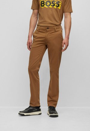 Pantalones ajustados marrones, hechos de una tela ligera, con cremallera, bolsillos frontales y un brillo sutil, combinados con zapatillas negras.