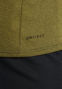 T-shirt vert olive en Dri-FIT avec une texture lisse, présentant un logo noir à l'ourlet, associé à un tissu noir en dessous.