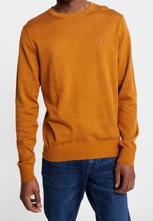 Mand iført en sennepsgul sweater med rund hals og et lille logo på brystet samt blå jeans, stående foran en ensfarvet baggrund.