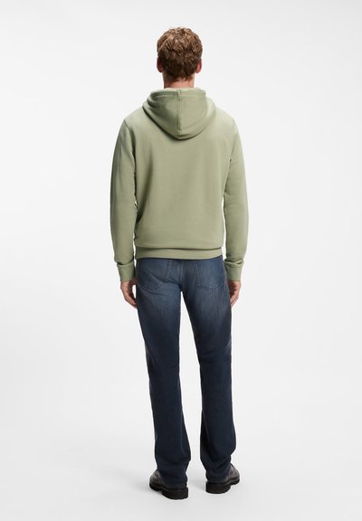 Homme debout de dos portant un sweat à capuche vert clair, un jean bleu foncé et des chaussures noires sur fond blanc uni.