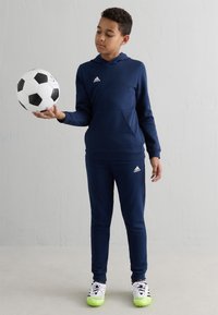 Ragazzo in tuta Adidas blu che bilancia un pallone da calcio bianco e nero sulla mano destra contro un muro leggero e semplice.