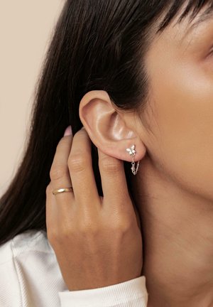 Silver butterfly earrings with a chain accent, featuring small clear gemstones, worn on a model's ear with a simple ring on her finger.