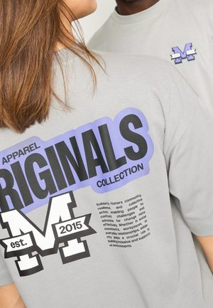 Dwie osoby ubrane w jasnoszare koszulki z nadrukiem "ORIGINALS", "M est. 2015" oraz tekstami o tematyce solidarności, w czarnym i fioletowym kolorze.