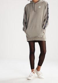 Felpa Puma grigia chiara, con tasca a marsupio, cappuccio con lacci e logo. Abbinata a leggings neri e sneakers bianche.