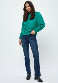 Felpa color teal con maniche a sbuffo, abbinata a jeans blu scuro e stivali neri alla caviglia. Design semplice e texture liscia.