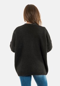 Mujer con cabello largo castaño, vistiendo un suéter negro holgado y jeans azules, de pie de espaldas contra un fondo blanco liso.