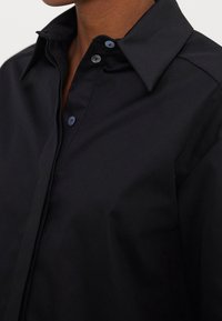 Camicia nera con bottoni, realizzata in tessuto liscio, dotata di colletto aperto e bottoni abbinati, con un focus su linee pulite e vestibilità su misura.