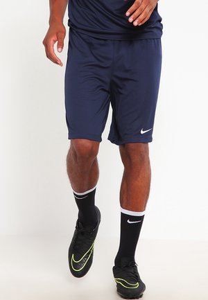Mann trägt dunkelblaue Nike-Shorts, schwarze Nike-Sportsocken und schwarze Fußballschuhe mit gelben Nike-Swoosh-Akzenten.
