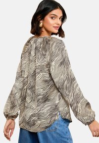 Long-sleeve blouse in cream with black wavy stripes, featuring a gathered neckline, elastic cuffs, and a slightly curved hem.