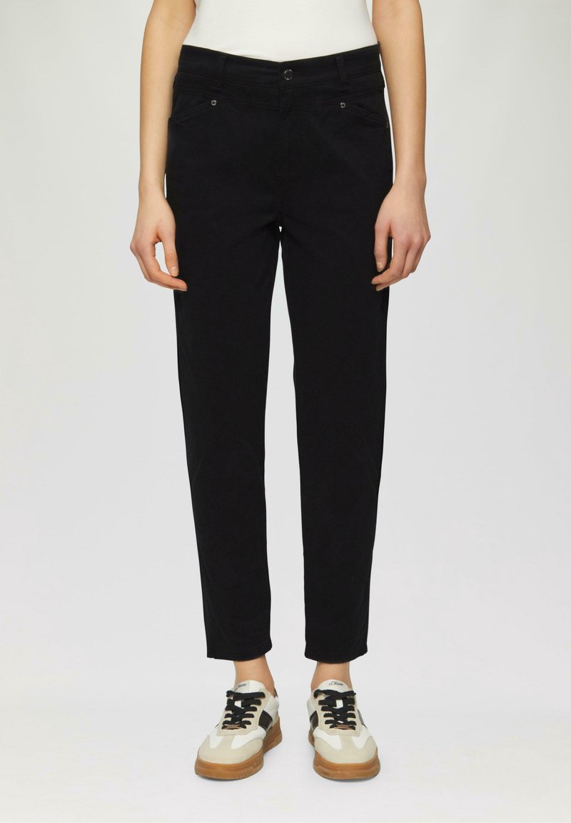 Personne portant un pantalon noir à jambes droites avec des poches avant, associé à des baskets blanches et beiges sur un fond gris clair.