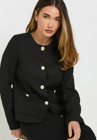 Mustapintainen tekstiilitakki, jossa on pyöreä kaula-aukko, kultaiset nappikiinnitykset ja kaksi etutaskua. Suunniteltu muotoonommelluksi ja pitkähihaiseksi.