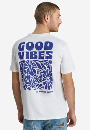 Uomo che indossa una T-shirt bianca con testo blu "GOOD VIBES" e design floreale sul retro, abbinata a jeans denim azzurri.