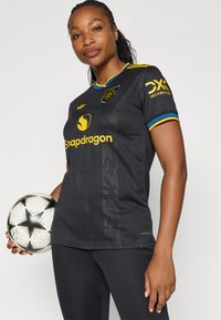 Melna sporta krekls ar dzelteniem un ziliem akcentiem, ar apaļu apkakli, logo un sponsora nosaukumu. To valkā persona, kas tur futbola bumbu.