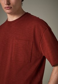 Tricou roșu din bumbac, cu decolteu rotund, mâneci scurte și buzunar la piept. Materialul are o textură subtilă și un croi lejer.