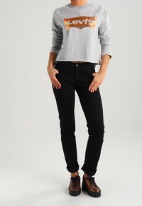 Sweatshirt gris court avec un logo Levi's orange usé, associé à un pantalon noir slim et des baskets brunes à lacets.