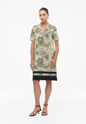 Femme portant une robe courte à manches courtes imprimée léopard avec un ourlet noir et à motifs, associée à des talons noirs à bride de cheville, debout en position neutre.