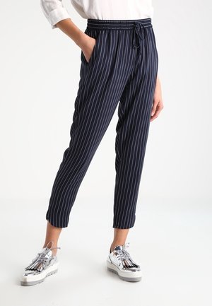 Person trägt marineblaue, gestreifte, gekürzte Hose mit elastischem Bund und weiße metallische Plateauschuhe, steht mit einer Hand in der Tasche.