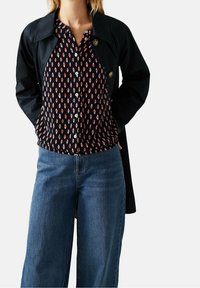 Cappotto nero sopra una blusa a fantasia blu navy con forme a diamante rosse e bianche, abbinato a jeans a gamba larga blu.