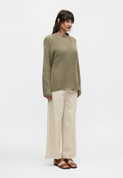 Γυναίκα που στέκεται, φοράει ένα πουλόβερ σε πράσινο ελιάς πλέξη, κρεμ παντελόνια με φαρδιά πόδια και καφέ πέδιλα με ανοιχτά δάχτυλα, μπροστά σε απλό λευκό φόντο.