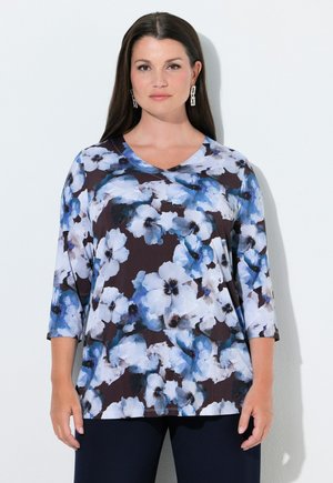 Frau mit langen dunklen Haaren, die ein schwarz-blaues, florales Dreiviertelarmoberteil und dunkle Hosen trägt, steht vor einem weißen Hintergrund.