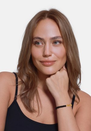 Bracciale di perle nere con un accento in catena dorata. Il modello indossa una maglia nera, con la mano appoggiata sulla guancia e i capelli che incorniciano dolcemente il viso.