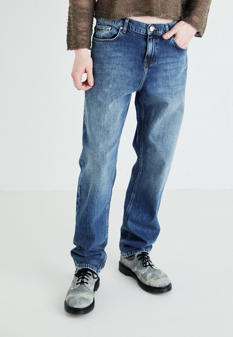Blå denimjeans med rak bendesign, som har en klassisk femficksstil och knappgylf. Bärs med grå snörskor.